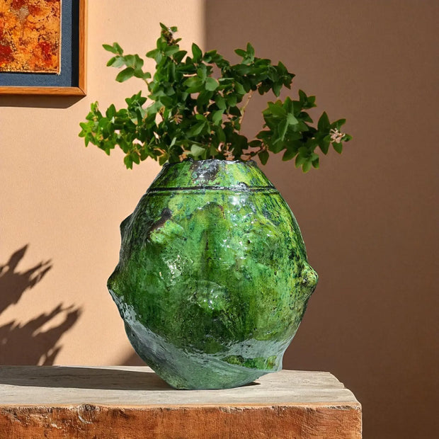 Green textured vase with plant on a wooden surface against a brown wall
