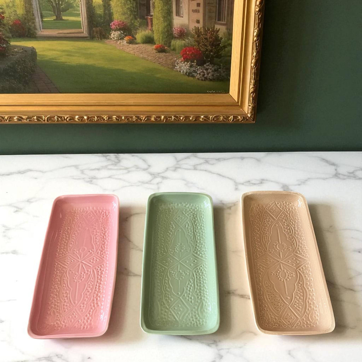 Three rectangular ceramic trays in pink, green, and beige on a marble surface with a painting in the background.