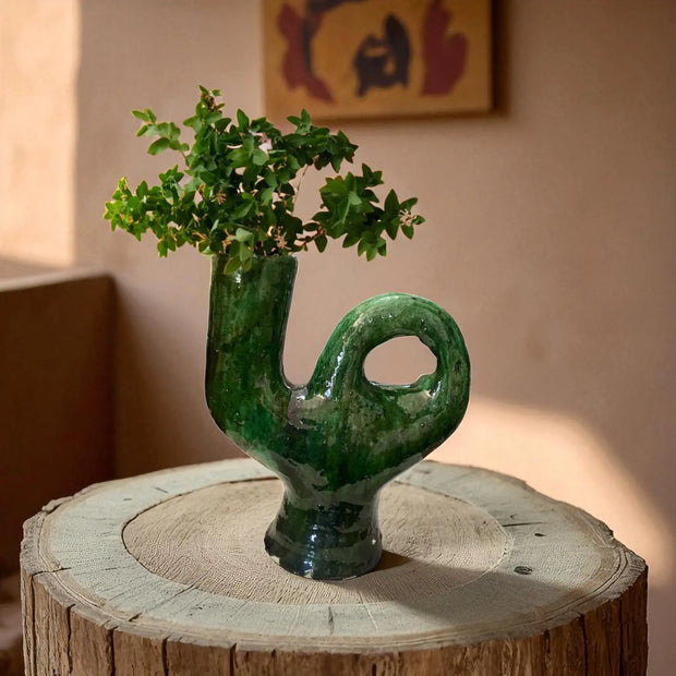 Green ceramic vase with plant on a wooden stump against a neutral wall.