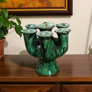 Green ceramic vase with black accents on a wooden surface, with a plant and framed picture in the background.