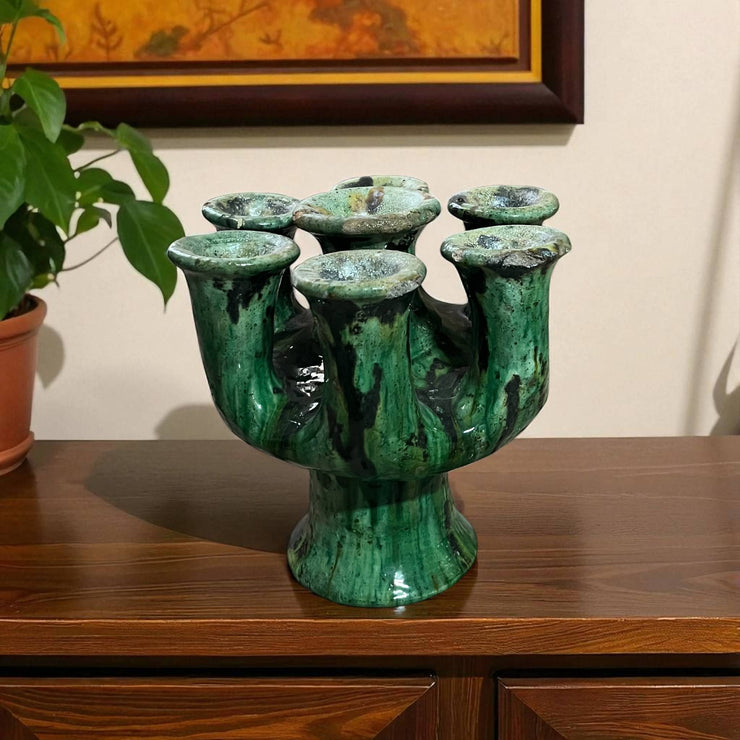 Green ceramic vase with black accents on a wooden surface, with a plant and framed picture in the background.