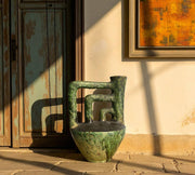 Vintage-style green ceramic vase on a wooden floor with a rustic wall and abstract painting in the background.