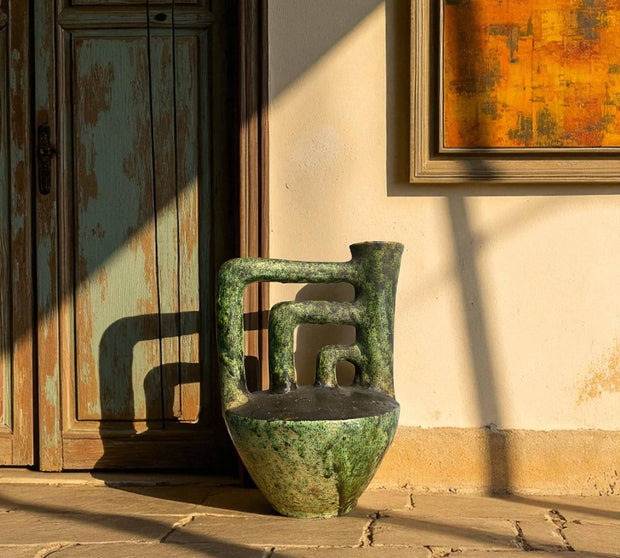 Vintage-style green ceramic vase on a wooden floor with a rustic wall and abstract painting in the background.