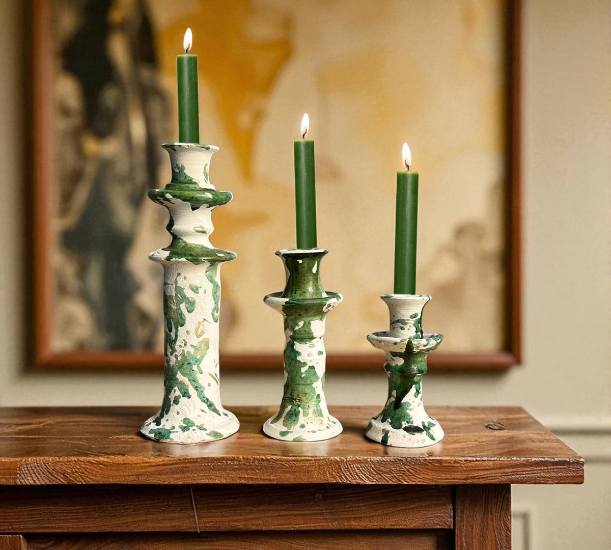 Three green candles in decorative ceramic candle holders on a wooden surface with a blurred painting in the background.