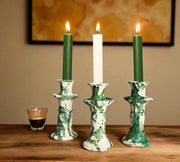 Three candlesticks with lit candles on a wooden surface, with a glass of coffee in the background.