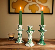 Three green candles in decorative candlesticks on a wooden surface with a glass of coffee and a painting in the background.