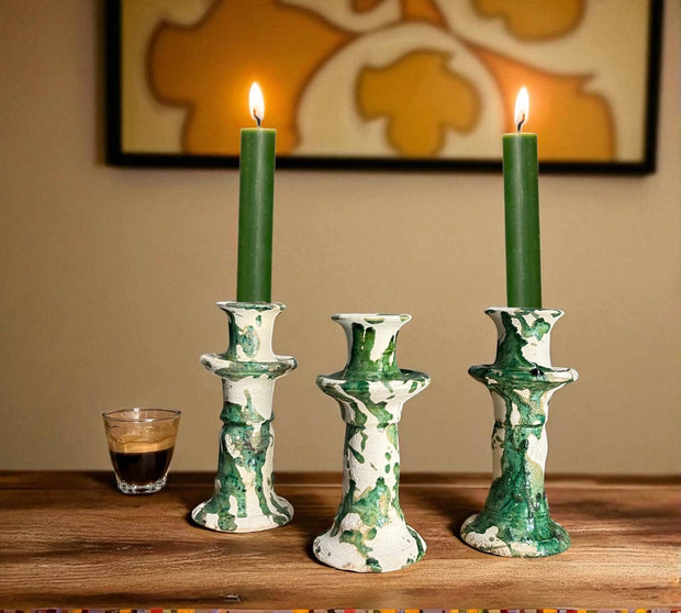 Three green candles in decorative candlesticks on a wooden surface with a glass of coffee and a painting in the background.
