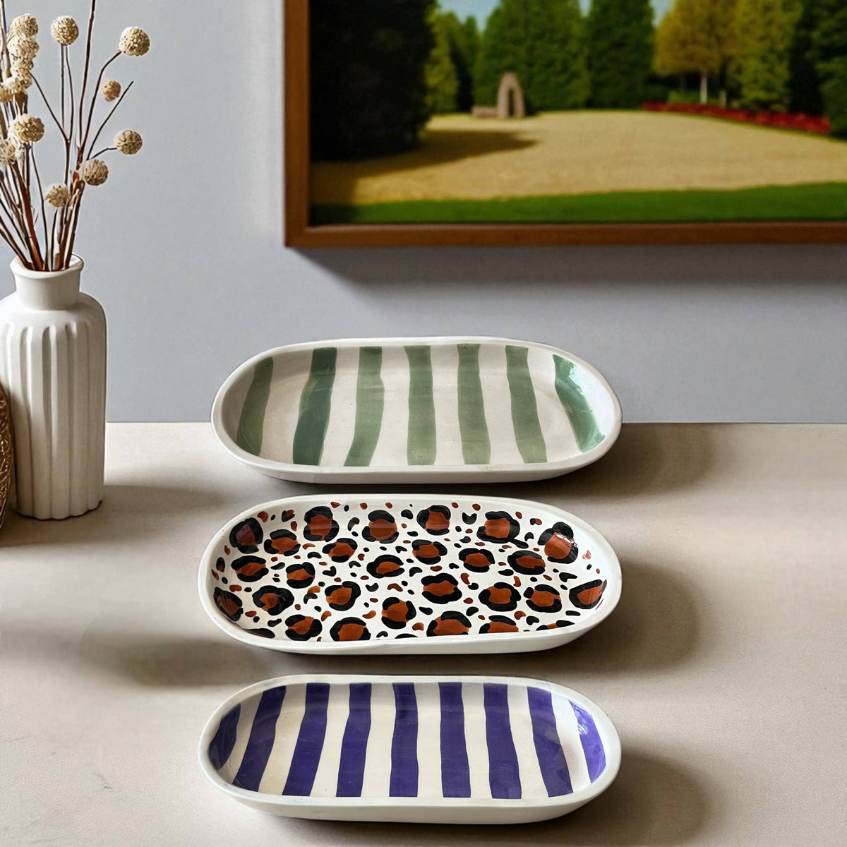 Three ceramic trays with different patterns on a surface, with a vase and framed picture in the background.