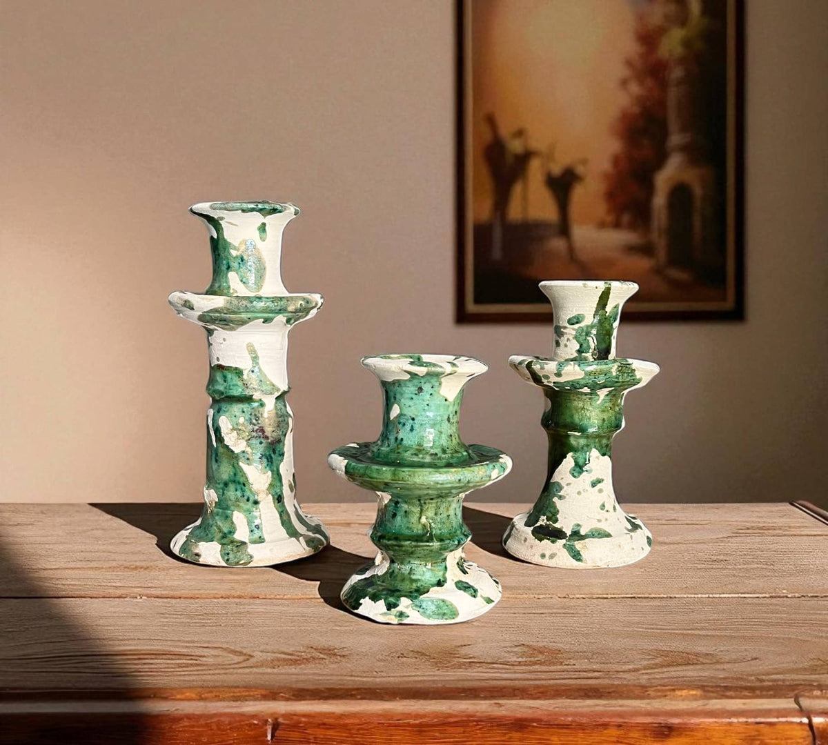 Set of three green and white ceramic candle holders on a wooden surface with a painting in the background.