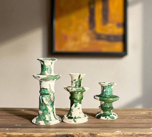 Three green and white ceramic candle holders on a wooden surface with a blurred abstract painting in the background.