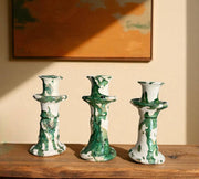 Three green and white ceramic candle holders on a wooden surface with a painting in the background.
