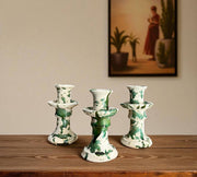 Three ceramic candle holders with green patterns on a wooden surface, with a painting in the background.