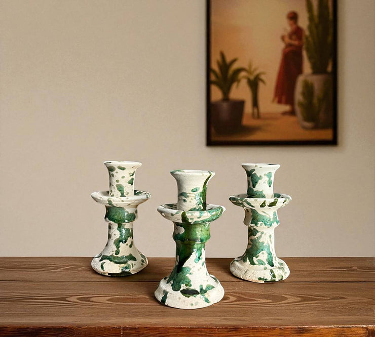 Three ceramic candle holders with green patterns on a wooden surface, with a painting in the background.