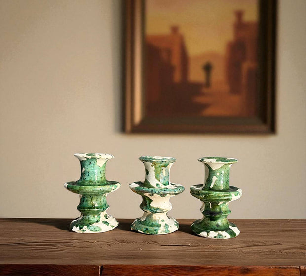 Three green ceramic candle holders on a wooden surface with a painting in the background.