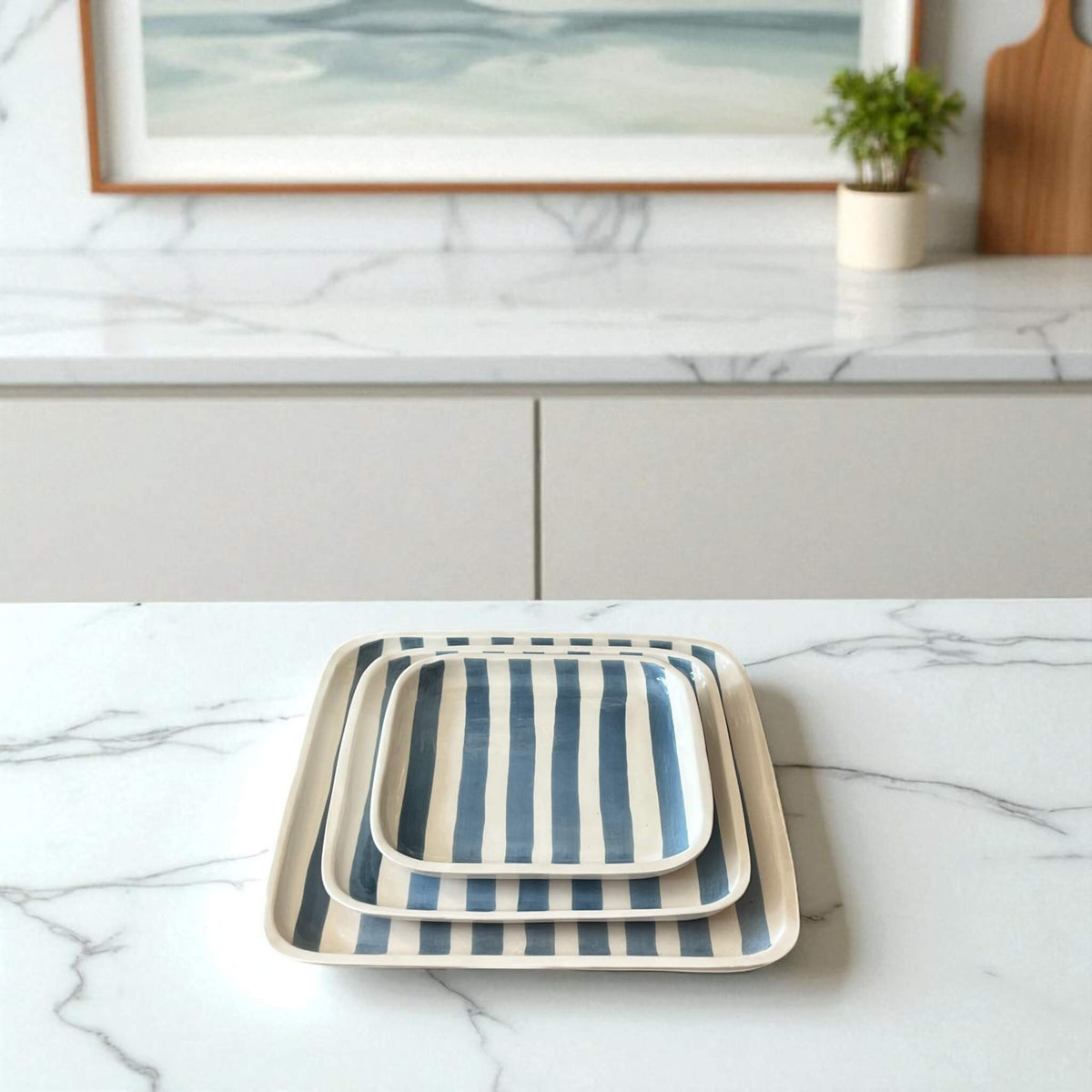 Set of three striped trays on a marble countertop with a framed picture and plant in the background.
