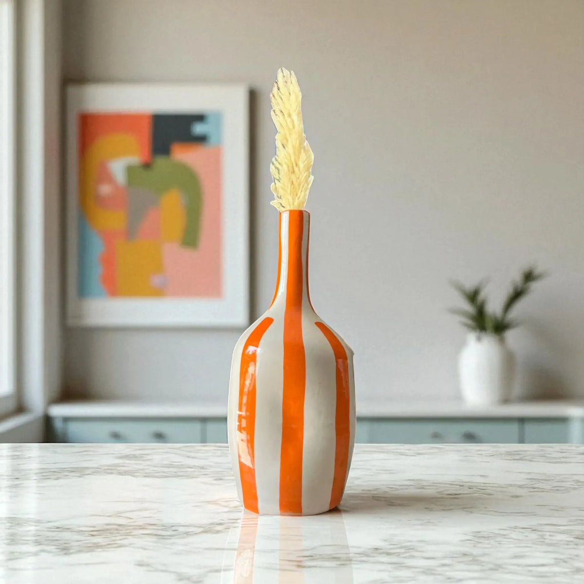 Orange and white striped vase with a decorative plant on a marble surface, with a blurred background.