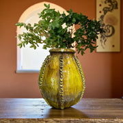 Decorative yellow vase with green branches on a wooden surface against an orange wall.