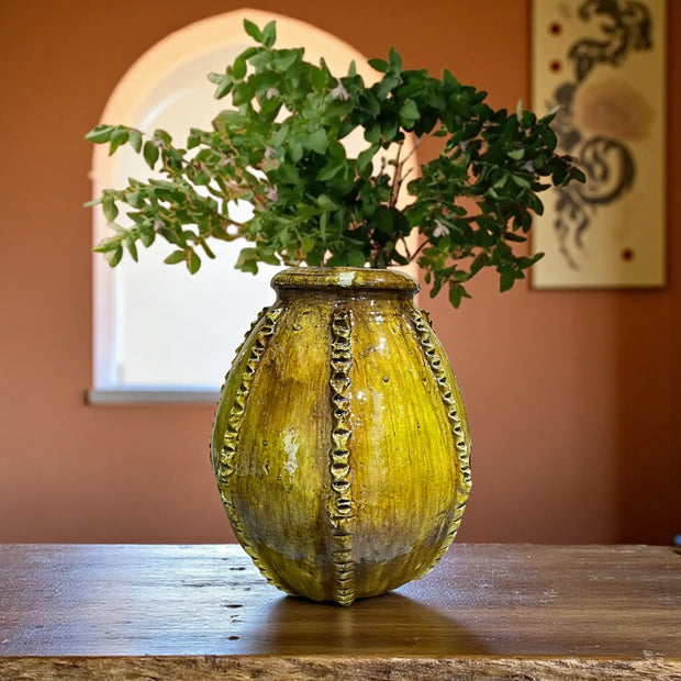 Decorative yellow vase with green branches on a wooden surface against an orange wall.