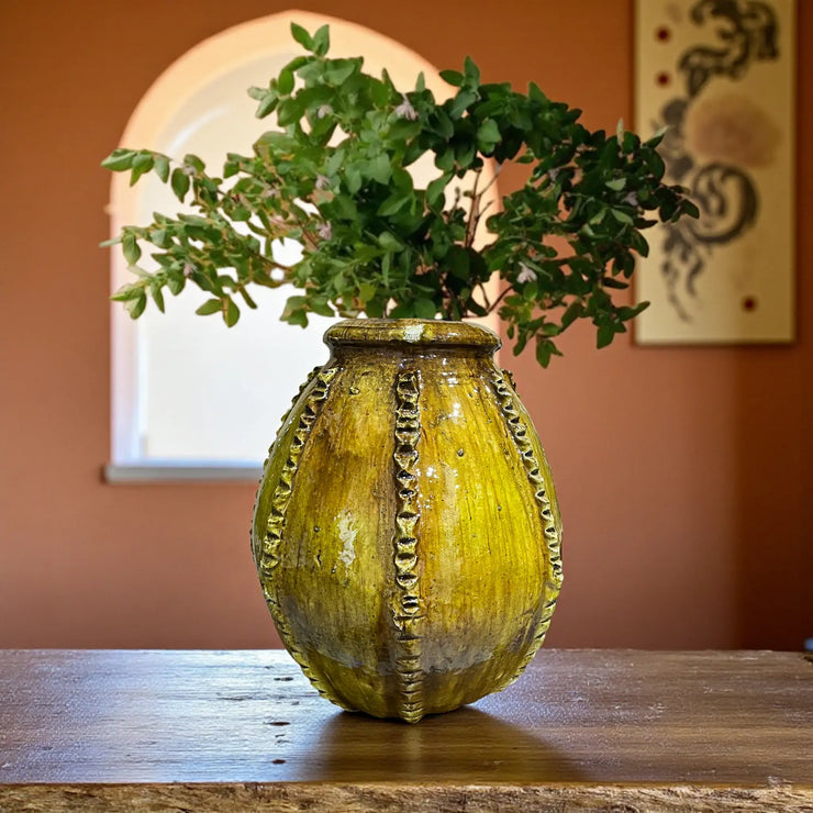 Decorative yellow vase with green branches on a wooden surface against an orange wall.