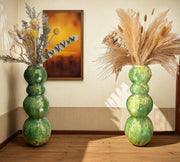 Two green ceramic vases with dried plants against a wall with a painting.