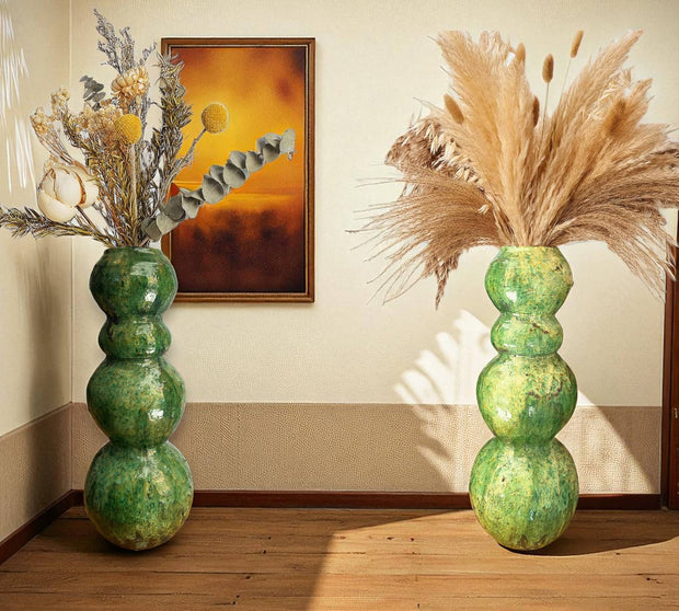Two green ceramic vases with dried plants against a wall with a painting.