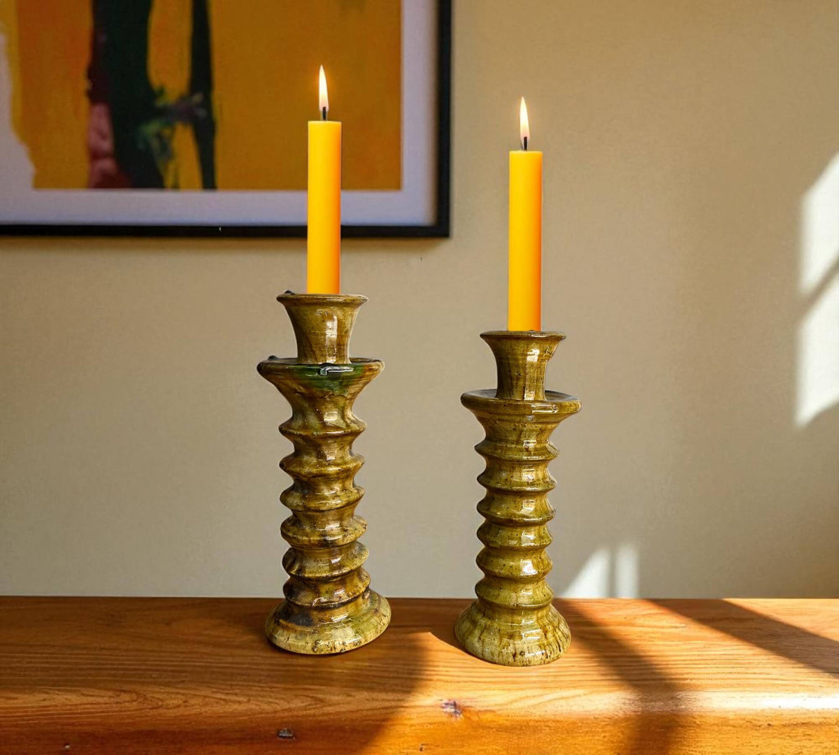 Two lit yellow candles in decorative green candle holders on a wooden surface with a neutral wall background.