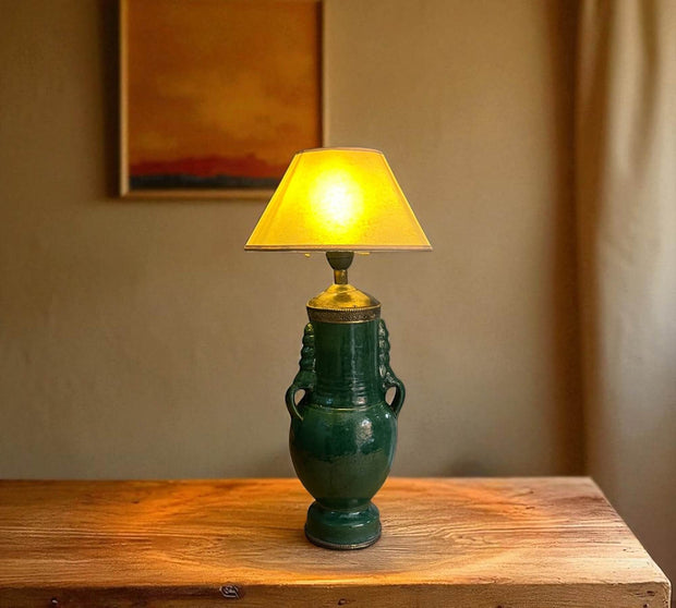 Green ceramic lamp with a yellow lampshade on a wooden surface against a beige wall.