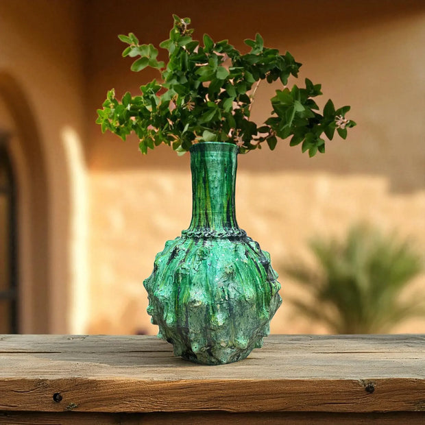 Green textured vase with plant on a wooden surface against a blurred outdoor background