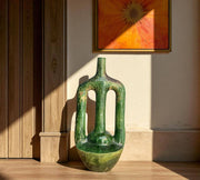 Green ceramic vase on a wooden surface with a wall painting in the background