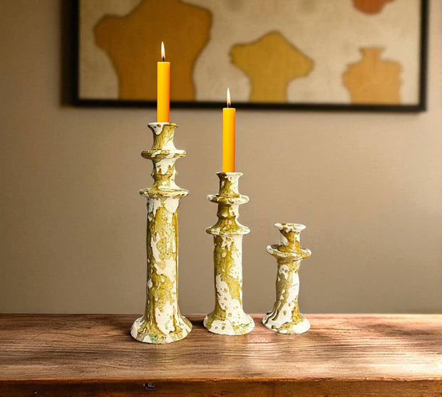 Three decorative candlesticks with candles on a wooden surface, framed artwork in the background.