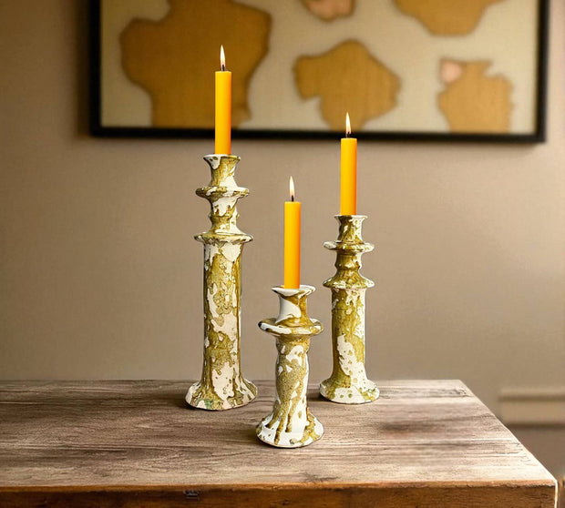 Three decorative candlesticks with lit candles on a wooden surface, with a neutral wall and abstract art in the background.
