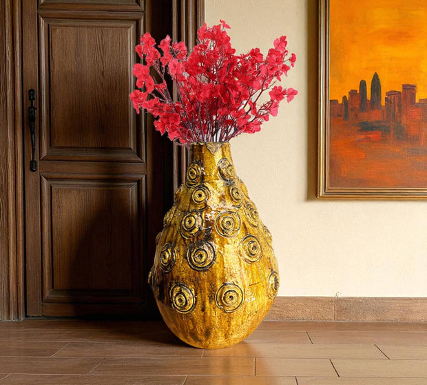 Decorative vase with gold circular patterns and red flowers in a room with wooden floor and wall art.