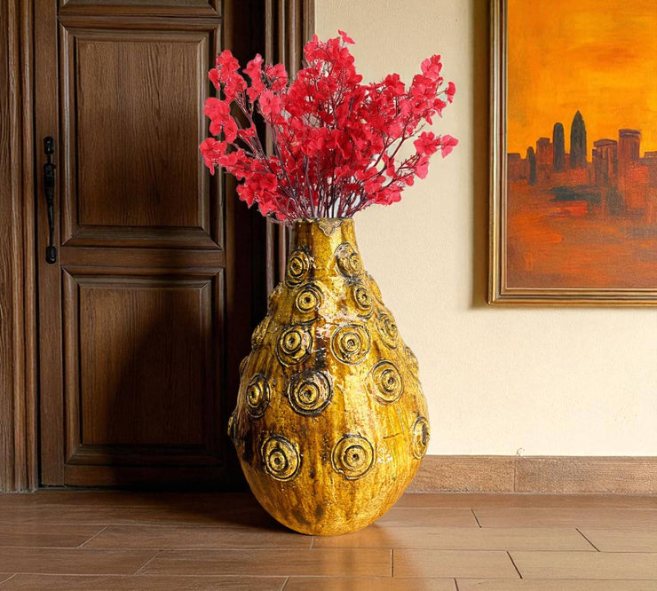 Decorative vase with gold circular patterns and red flowers in a room with wooden floor and wall art.