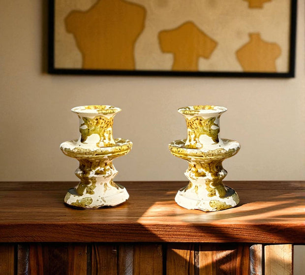 Two gold and white decorative candle holders on a wooden surface with a framed abstract painting in the background.