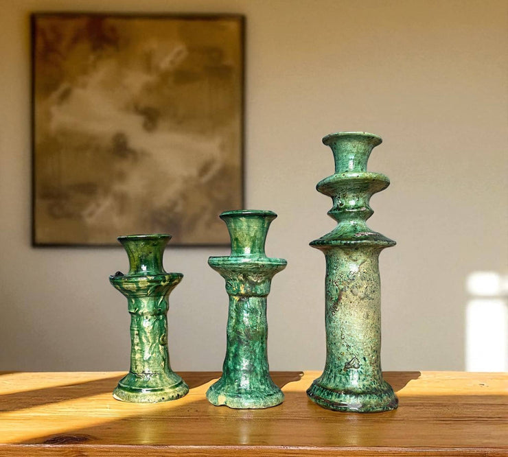 Three green ceramic candle holders on a wooden surface with a blurred painting in the background.
