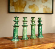 Four green ceramic candle holders on a wooden surface with a blurred painting in the background.