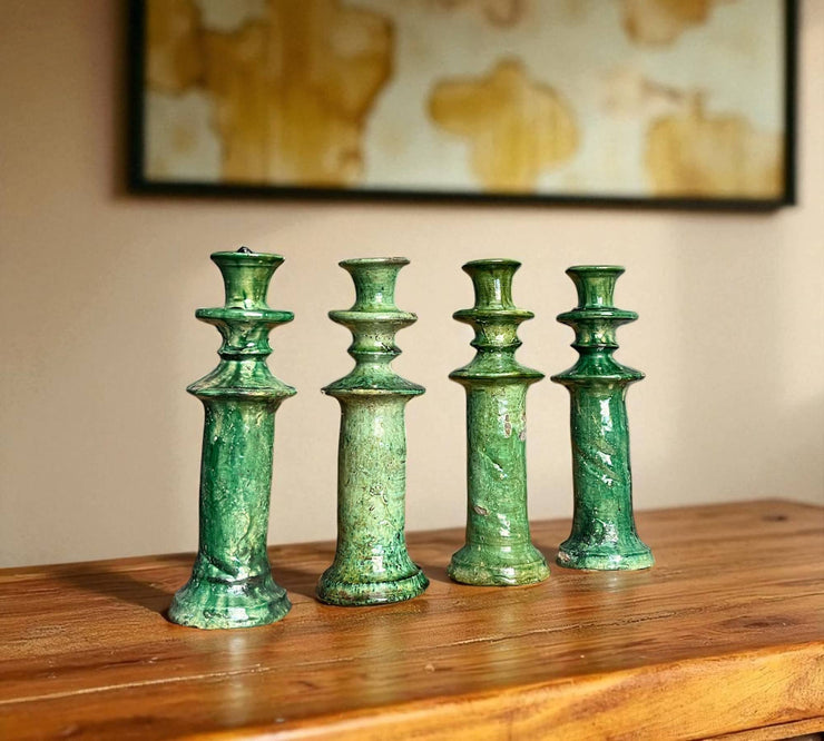 Four green ceramic candle holders on a wooden surface with a blurred painting in the background.