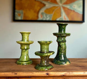 Three green ceramic candle holders on a wooden surface with a colorful abstract painting in the background.