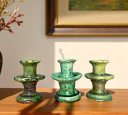 Three green ceramic candle holders on a wooden surface with a painting in the background.