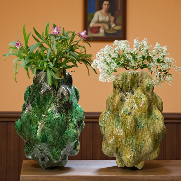 Two unique, textured vases with plants on a wooden surface against an orange wall.