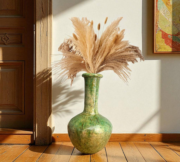 Green vase with dried pampas grass against a wooden floor and wall.