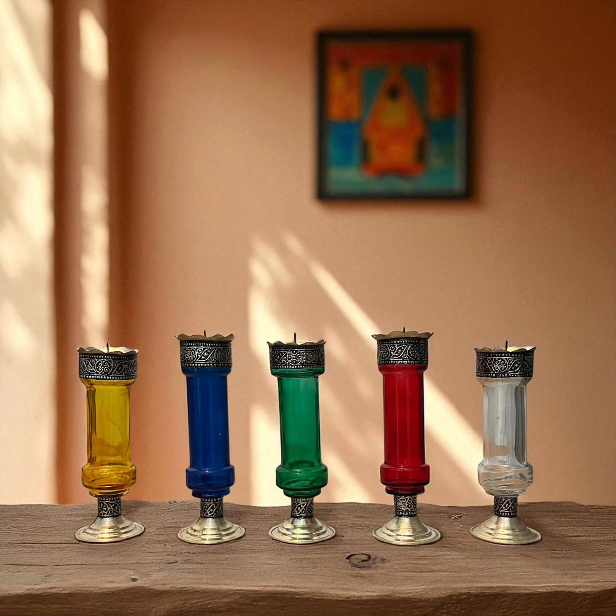 Five Vintage hand‑engraved silver‑plated brass and recycled glass candlesticks in assorted colours on a wooden surface with a blurred painting in the background.