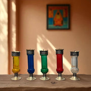 Five Vintage hand‑engraved silver‑plated brass and recycled glass candlesticks in assorted colours on a wooden surface with a blurred painting in the background.