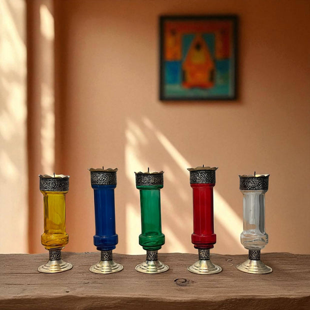 Five Vintage hand‑engraved silver‑plated brass and recycled glass candlesticks in assorted colours on a wooden surface with a blurred painting in the background.