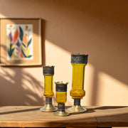 Three yellow Vintage hand‑engraved silver‑plated brass and recycled glass candlesticks on a wooden surface, framed artwork in the background.