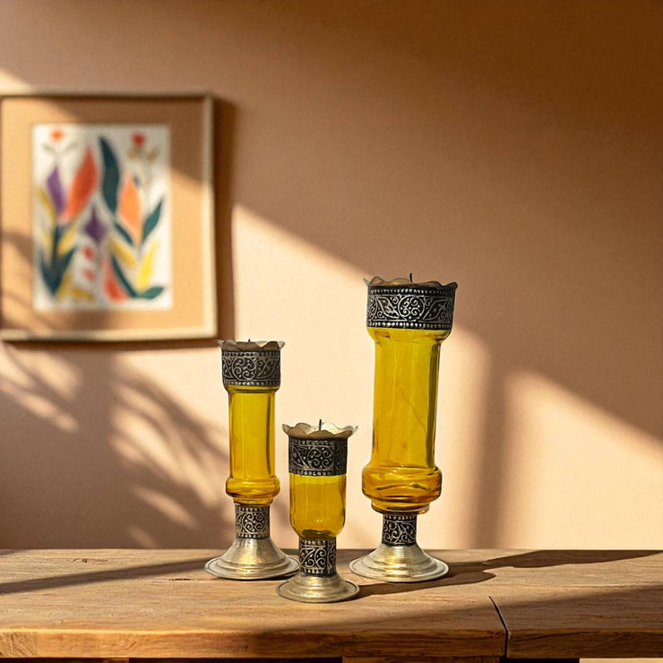 Three yellow Vintage hand‑engraved silver‑plated brass and recycled glass candlesticks on a wooden surface, framed artwork in the background.