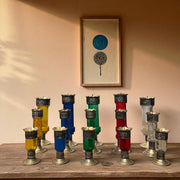 Vintage hand‑engraved silver‑plated brass and recycled glass candlesticks in assorted colours on a wooden surface with a framed artwork in the background.