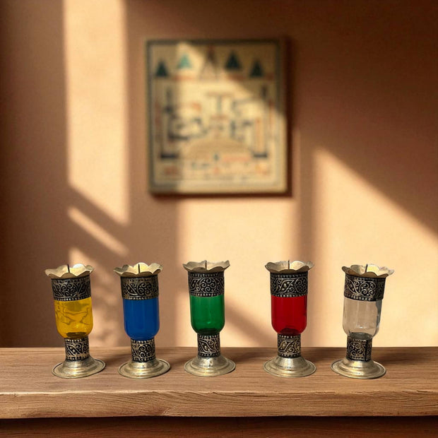 Vintage hand‑engraved silver‑plated brass and recycled glass candlesticks in assorted colours on a wooden surface with a wall painting in the background.