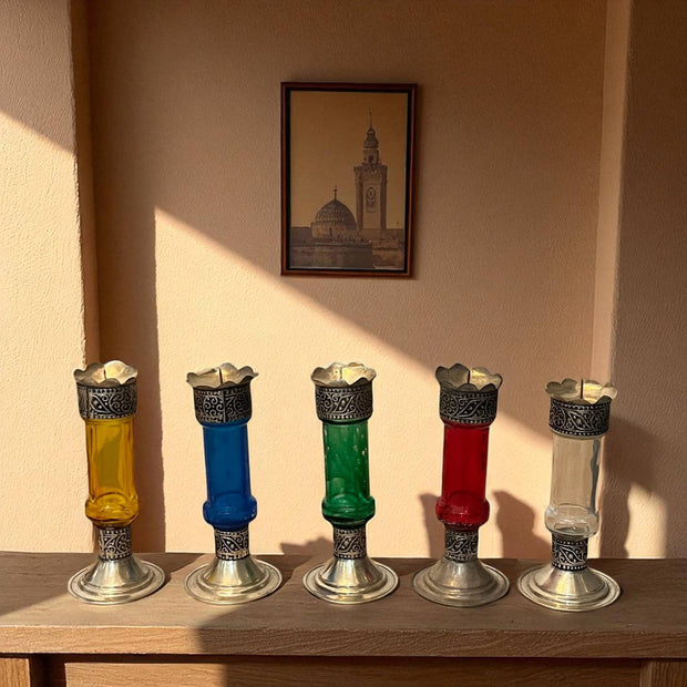 Five Vintage hand‑engraved silver‑plated brass and recycled glass candlesticks in assorted colours on a wooden surface with a framed picture of a tower in the background.