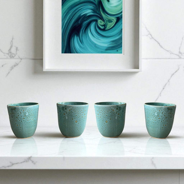 Four teal ceramic cups on a marble surface with a abstract teal painting in the background.